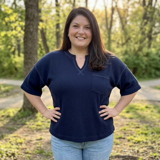 A woman stands outdoors in a wooded park setting wearing a dark navy blue ribbed short-sleeve henley top with a chest pocket, paired with light blue jeans.